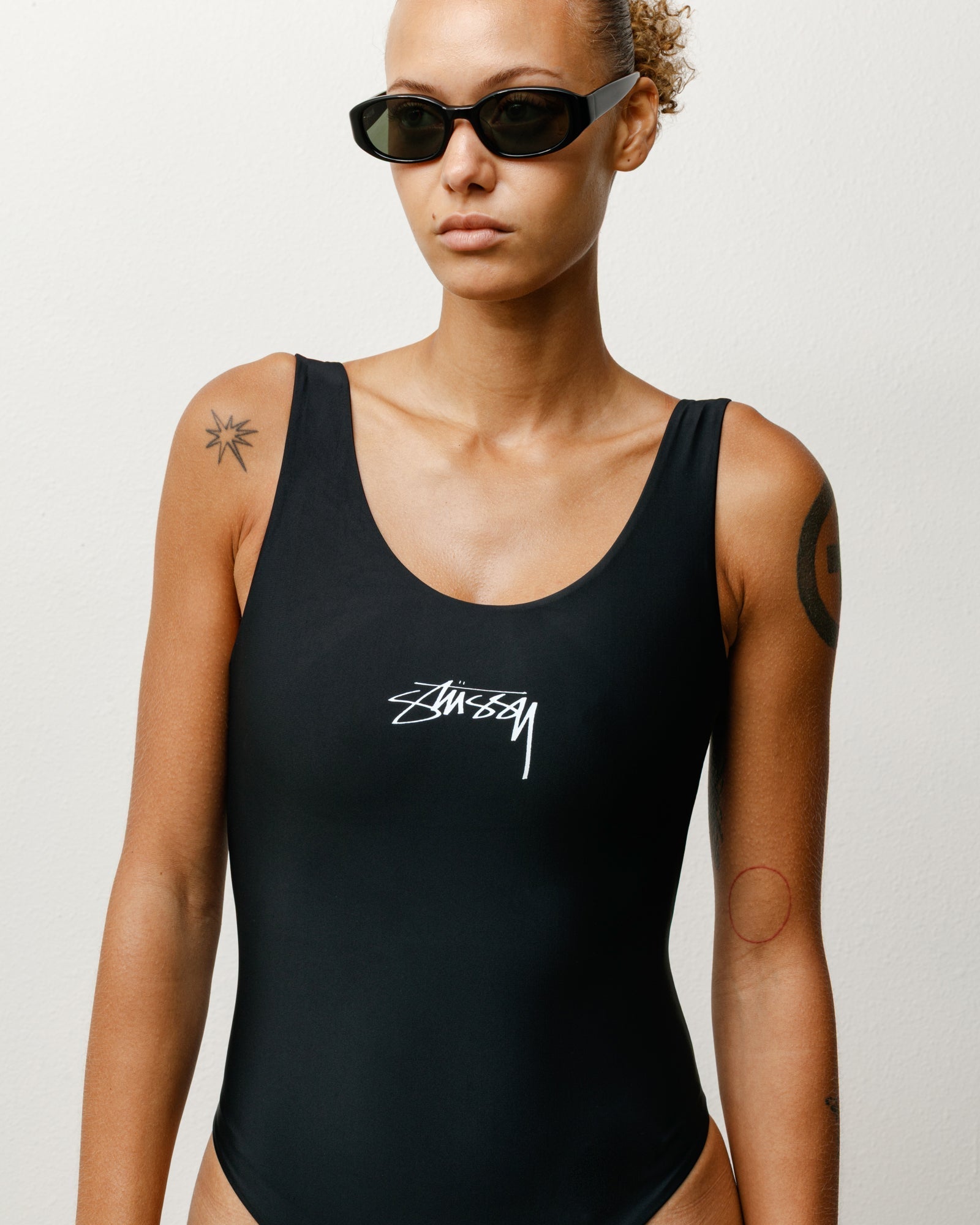 胸元にStüssyのストックロゴをプリントしたブラックのSurf Stock One Piece Swimsuitを着用し、サングラスを合わせた女性モデルのバストアップショット。