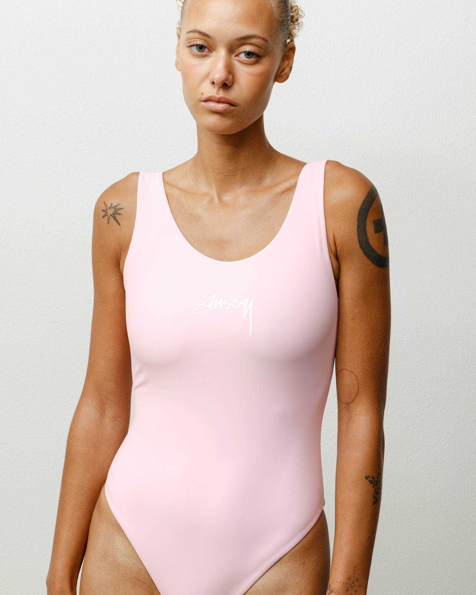 Surf Stock One Piece ウィメンズ レギュラー イン ピンク。Stüssyのロゴが配された、スクープネックとハイカットレッグが特徴的なフィット感のあるデザインのスイムウェア。