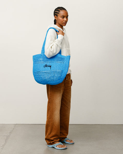ブルーのラフィア素材で編み上げたStüssyストックロゴ刺繍入りのWoven Tote Bag アクセサリーを肩に掛けたスタイリング。