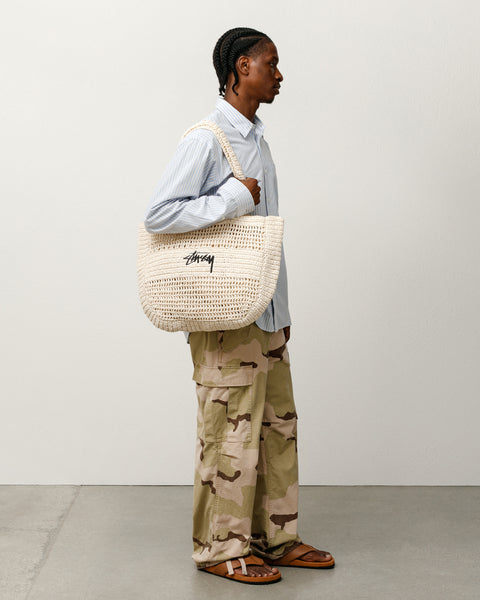 アイボリーのStüssy ウーブン トートバッグ アクセサリー。ラフィア素材に黒のストックロゴ刺繍をあしらったハンドメイドのバッグを、モデルが肩に掛けている着用シーンです。