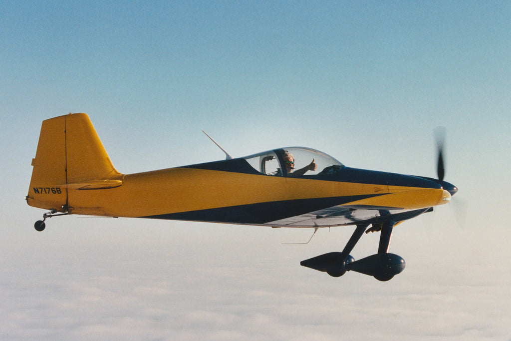 A small, yellow and dark blue propeller airplane flying high in the sky above a layer of clouds, with the pilot wearing the Adrian Sunglasses in Frost Grey/Green Mirror Lens.