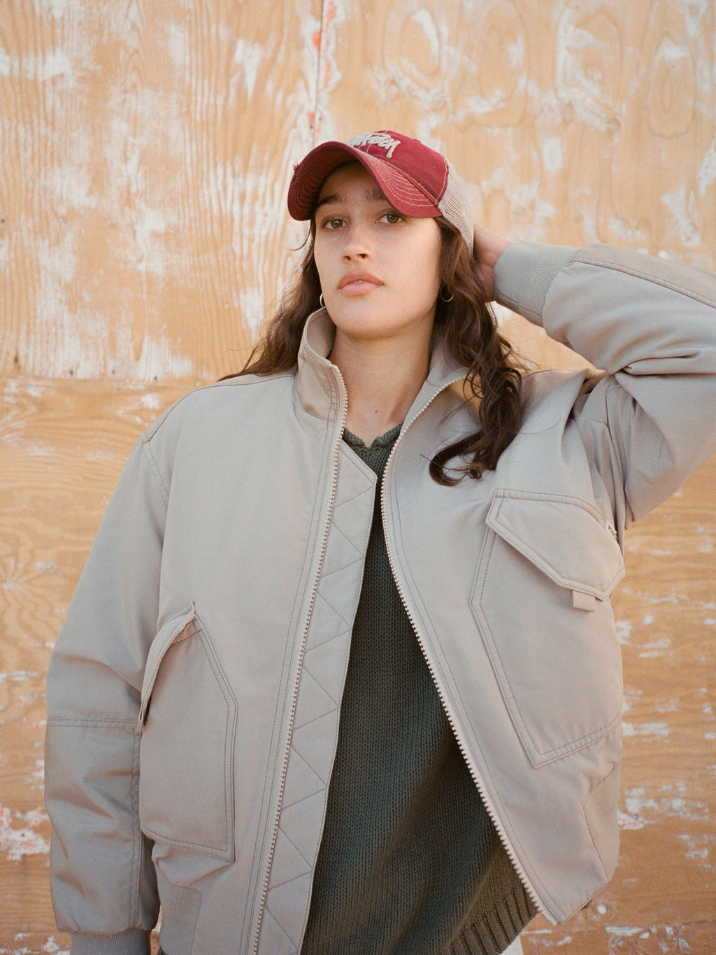 Person wearing the New Era 9Twenty Washed Basic Trucker in Maroon and Mock Neck Flight Bomber in Khaki over the Roll V Neck Sweater in Olive posing in natural sunlight against a textured plywood wall.