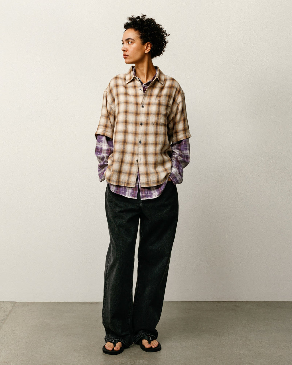 Person wearing the Liam SS Plaid Shirt in Brown layered over a long sleeve plaid shirt and dark pants against a plain background.