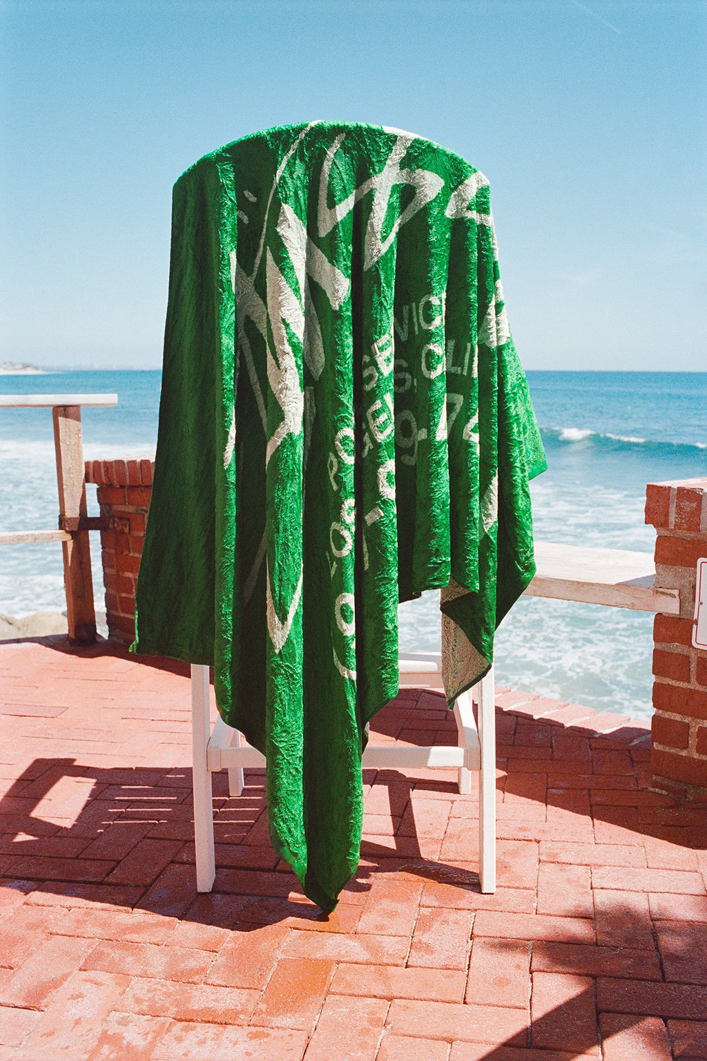 The Pool Service Beach Towel in Green draped over a chair on a brick patio overlooking the ocean.