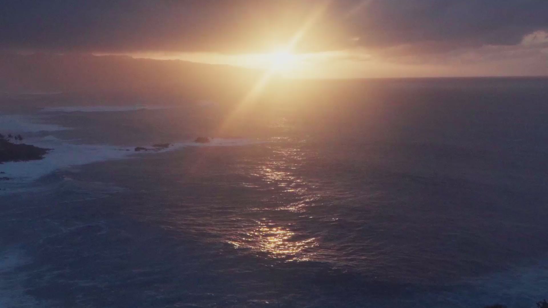 Golden sunlight breaks through dark clouds over the ocean at sunrise or sunset, casting a bright reflection across the water while waves roll gently toward a rocky shoreline.