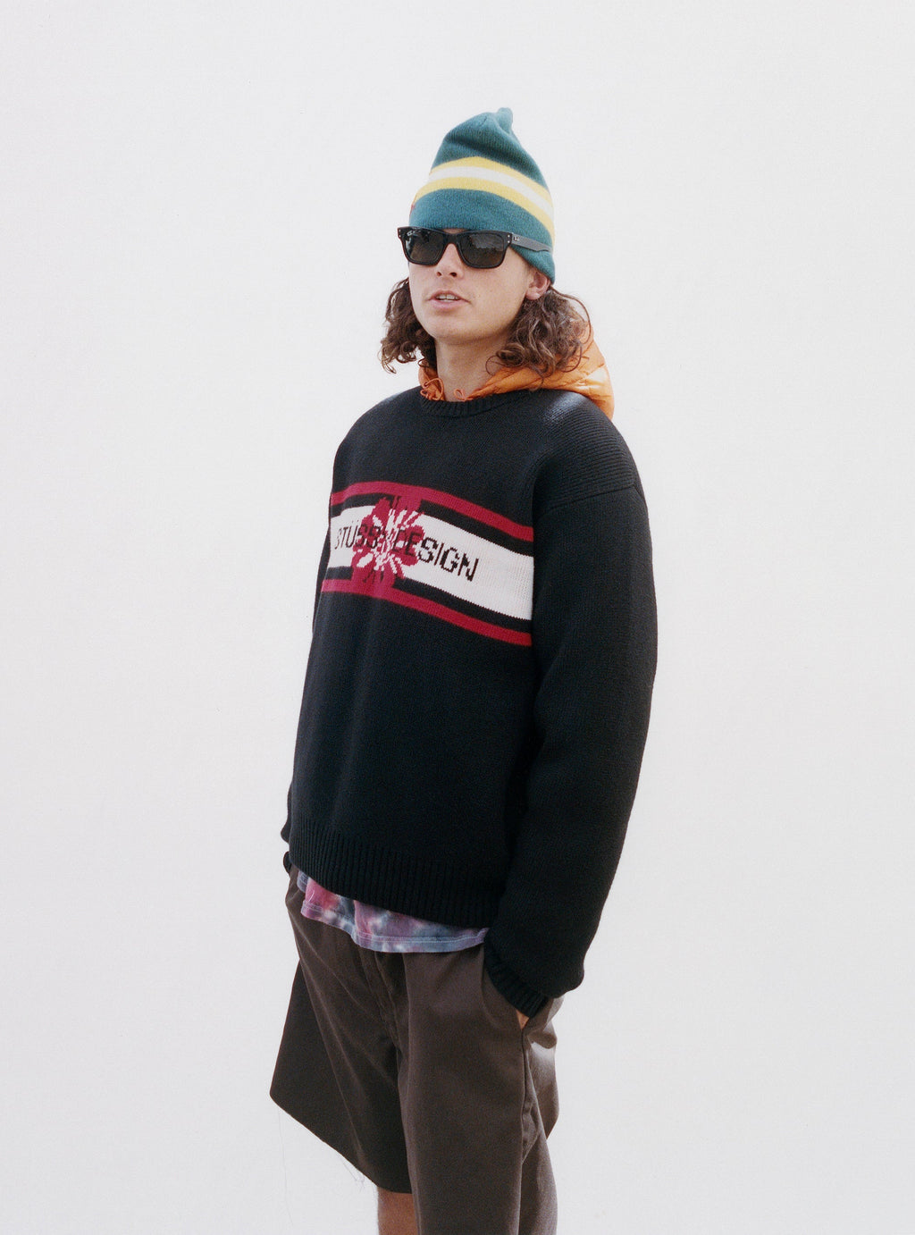 A man with long curly hair wearing sunglasses, a colorful beanie, and the STRIPED HIBISCUS SWEATER in BLACK posing against a white background.