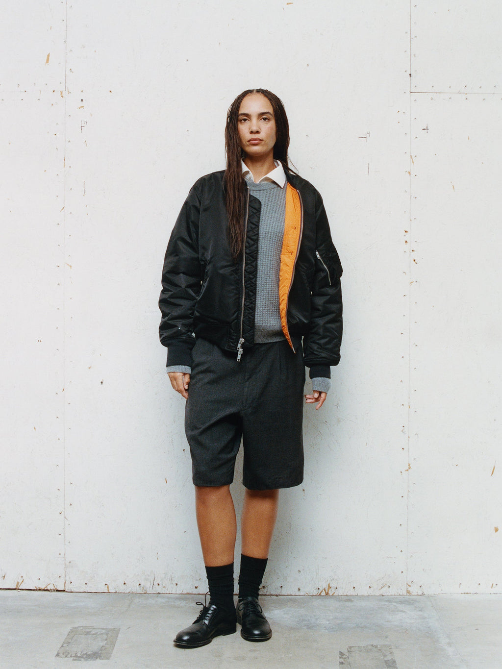 Full-length portrait of a model standing against a textured white wall, wearing the MA-1 Bomber Jacket in Black layered over a grey crewneck sweater, a white collared shirt, dark grey knee-length shorts, black socks, and black leather dress shoes.