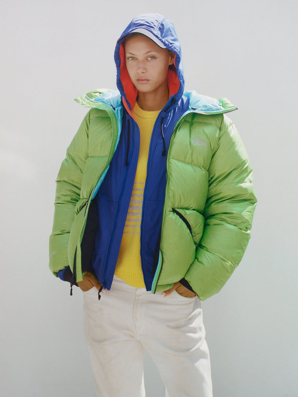 Model wears the Stüssy Down Parka Micro Ripstop in Kiwi over a blue hooded layer, yellow top and white pants.
